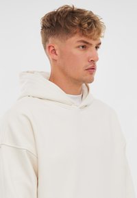 Hoodie de color beige claro con un corte relajado, que cuenta con una capucha con cordón, puños acanalados y una textura suave. Diseño minimalista sin patrones.