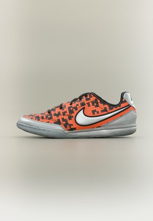 Lav sportssko med et lyst orange og sort pixeleret mønster, sølvfarvet sål, stort hvidt Nike-logo og sorte snørebånd på en neutral baggrund.