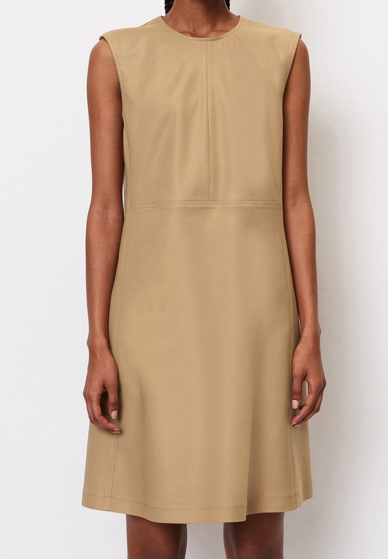 Robe beige sans manches longueur genou avec épaules structurées portée par une personne debout devant un fond uni.