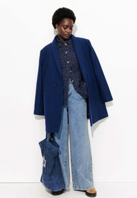 Blazer en laine bleu sur une chemise marine à motifs, associé à un jean large bleu clair et un sac fourre-tout bleu. Des bottes marron complètent la tenue.