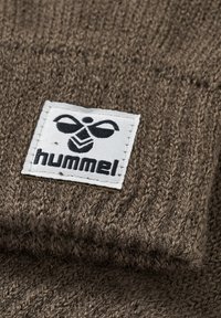 Un tissu tricoté marron présente une étiquette rectangulaire blanche avec le texte et le logo "hummel" en noir. La texture est douce avec des détails de couture visibles.