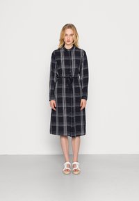 Vestido xadrez preto e branco com mangas longas, fecho de botão, cinto na cintura e comprimento até à meia-ақela, combinado com sandálias brancas.