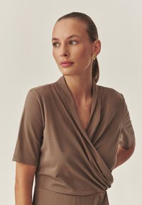 Braunes Wickelkleid mit kurzen Ärmeln, das eine glatte Textur und einen tiefen Ausschnitt aufweist. Das Modell zeigt eine Seitenansicht vor einem neutralen Hintergrund.