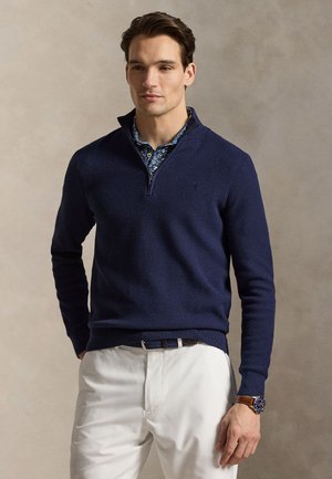 Férfi sötétkék texturált, negyed cipzáras pulóvert visel virágmintás ing felett, fehér nadrágot, barna övet és barna órát bézs háttér előtt.