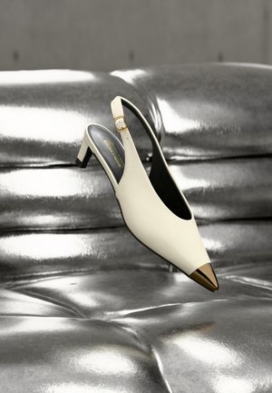 Escarpin blanc à bride arrière avec bout pointu doré, présenté sur une surface capitonnée métallique argentée brillante.