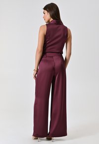 Top senza maniche in satin bordeaux abbinato a pantaloni a gamba larga. Presenta un colletto elegante e una texture liscia, con una vestibilità sartoriale in vita.