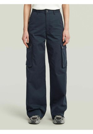 Pantaloni cargo blu navy alla caviglia con grandi tasche laterali, chiusura con bottoni, indossati con sneaker grigie e bianche e un top bianco visibile in vita.