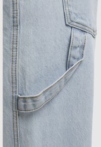 Zbliżenie jasnoniebieskiego denimu z boczną kieszenią i brązowymi szwami, ukazujące fakturę i detale materiału oraz szwów.
