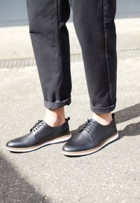 Chaussures en cuir noir à finition lisse, semelle en caoutchouc blanc et accents marron. Lacées avec des lacets foncés, adaptées pour un usage décontracté ou semi-formel.
