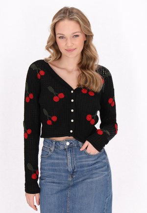 Cardigan nero corto con texture a costine, caratterizzato da ricami di ciliegie rosse e foglie verdi, e bottoni perla sul davanti. Abbinato a una gonna di jeans.