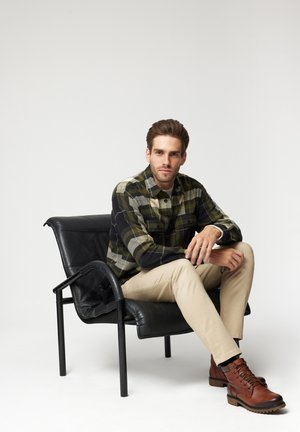 Koszula w kratę flanelowa w zielonym, czarnym i beżowym kolorze, w połączeniu z beżowymi spodniami i brązowymi skórzanymi butami, siedząca na czarnej skórzanej krześle.