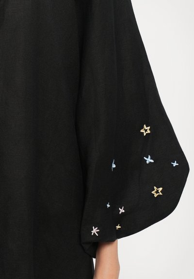 Zwarte stof top met brede vleermuismouwen, versierd met kleurrijke sterrenborduring in pastel tinten van blauw, roze en goud langs de randen.