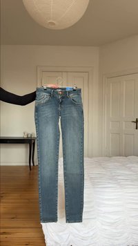 Személy kék farmernadrágot tart egy vállfán egy hálószobában, ahol fa padló és fehér ágytakaró van.