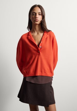 Mujer con un suéter naranja suelto con botones sobre una blusa marrón oscuro y una falda negra asimétrica sobre un fondo liso y claro.