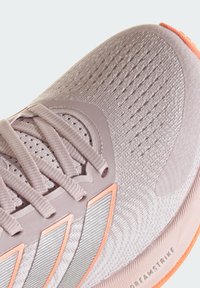 Scarpa sportiva di colore viola chiaro con texture a maglia, design con lacci e dettagli arancioni. Presenta sovrapposizioni in materiale sintetico e suola in gomma.