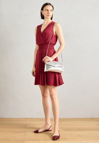 Robe sans manches bordeaux avec un décolleté en V et une taille froncée, accompagnée d'un sac à épaule métallique argenté et de chaussures à talons transparents.