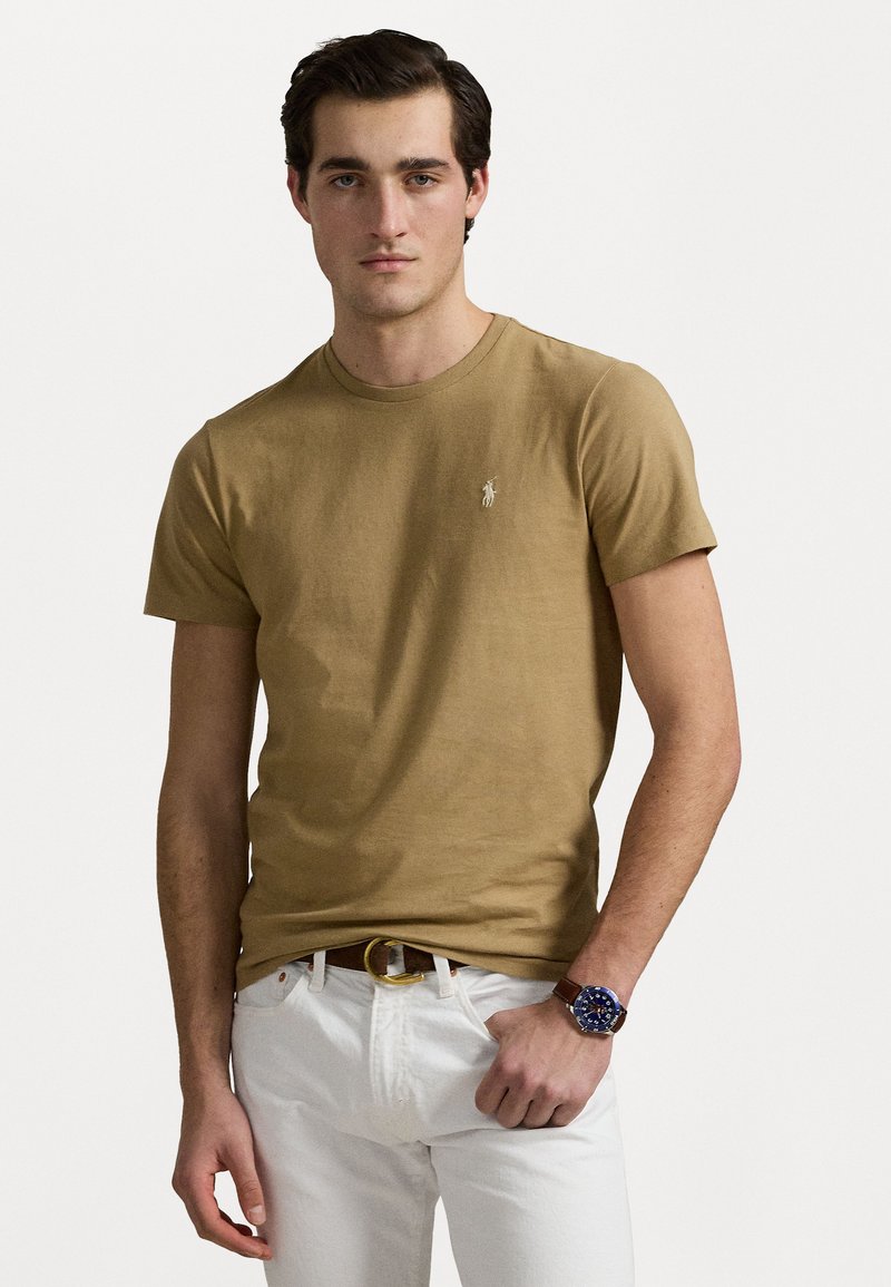 Tan cotton T-shirt with a crew neck and short sleeves, featuring a small embroidered logo on the chest, paired with white jeans and a brown belt.
