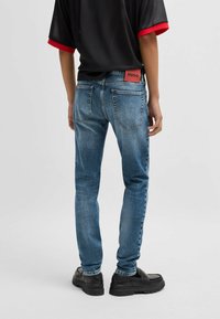 Jeans da uomo in denim blu, slim fit, con una finitura sbiadita. Presentano un'etichetta rossa con logo sul retro, tasche standard e dettagli di cucitura.