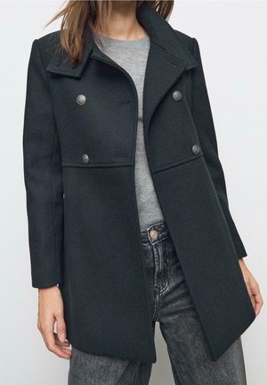 Femme portant un caban noir croisé sur un pull gris et un jean noir délavé, sur un fond uni.