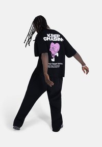 Svart oversized t-skjorte med en stor rosa grafikk av et hjerte med hender på baksiden, ledsaget av fet hvit tekst. Brukt med svarte bukser.