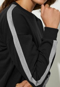 Donna che indossa una maglietta nera a maniche lunghe con strisce riflettenti argento che scendono lungo le maniche, con la mano appoggiata vicino al mento.