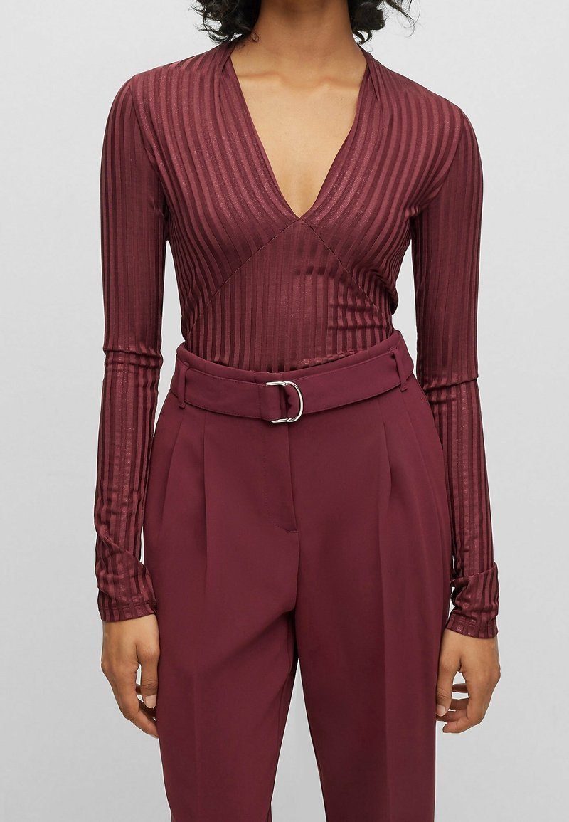 Personne portant un haut côtelé à manches longues avec un décolleté en V profond et un pantalon taille haute ceinturé de couleur bordeaux foncé assortie.