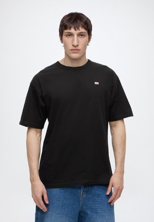 Jeune homme aux cheveux courts et foncés portant un t-shirt Diesel noir et un jean bleu, debout devant un fond blanc uni.