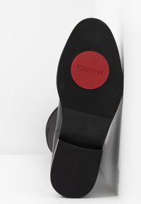 Bota negra con una suela de goma texturizada. Logo circular rojo en la suela y detalles adicionales en los lados. Material superior suave.