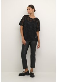 Schwarzes Spitzentop mit floralen Mustern, kombiniert mit eleganten schwarzen Kunstlederhosen und schwarzen Mokassins mit goldenen Akzenten.