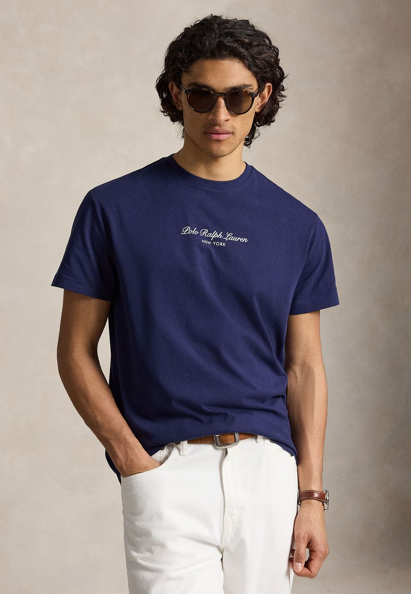 Άνδρας που φοράει σκούρα γυαλιά ηλίου, μπλε Polo Ralph Lauren t-shirt, λευκό παντελόνι με καφέ ζώνη και καφέ δερμάτινο ρολόι, στέκεται μπροστά σε απλό φόντο.