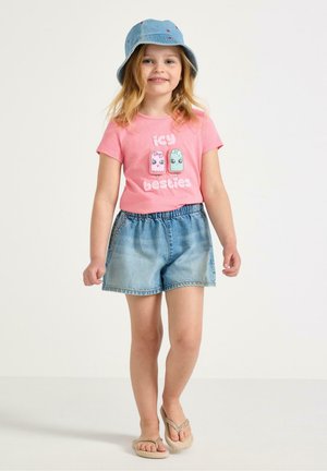 Jong meisje in een roze "icy besties" t-shirt, spijkershorts, blauwe bucket hat en beige slippers, staand en glimlachend tegen een witte achtergrond.