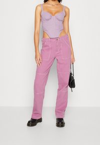 Kvinna som bär en lila korsett topp med utskärningar, rosa cargo byxor, svarta stövlar och håller en liten svart handväska med kedjehandtag.