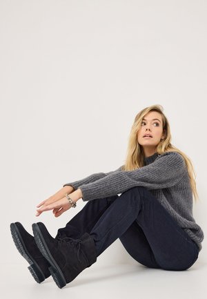 Bottes en suède noires à lacets avec des semelles en caoutchouc épaisses, associées à un pull en maille gris et un jean en denim foncé. Position assise avec les jambes étendues.