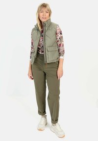 Gepolsterte olivgrüne Weste mit hohem Kragen, Reißverschluss vorne, zwei Taschen, getragen über einem blumigen Langarmhemd und olivfarbenen Hosen, kombiniert mit Turnschuhen.