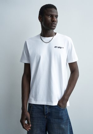 Νέος άντρας που φοράει λευκό t-shirt Off-White και μπλε τζιν, κοιτάζει προς τα πλάγια με το ένα χέρι στην τσέπη, μπροστά σε απλό ανοιχτό φόντο.