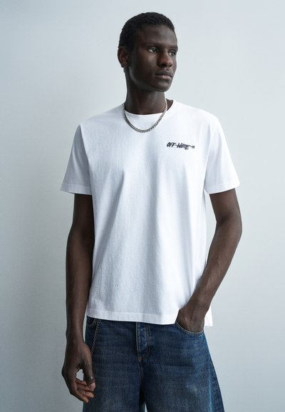 Jeune homme portant un t-shirt blanc Off-White et un jean bleu, regardant sur le côté avec une main dans la poche, sur un fond clair uni.