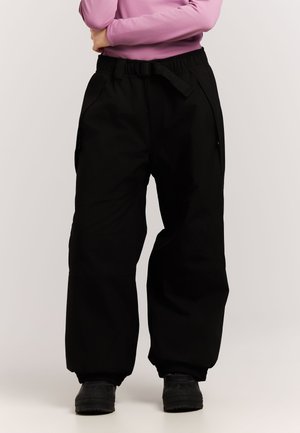 Personne portant un pantalon noir ample avec poignets élastiques et ceinture, associé à des chaussures noires et un haut rose à manches longues.
