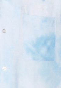 Maglietta tie-dye azzurro chiaro nella parte frontale con una tasca sul petto a destra e due bottoni visibili sul lato sinistro.