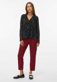 Mujer vestida con blusa negra de volantes con manchas blancas, pantalones burdeos hasta los tobillos y mocasines en blanco y negro, de pie contra un fondo blanco.