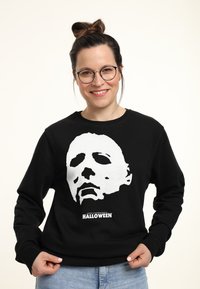 Felpa nera con una grafica bianca di un volto mascherato, con il testo "John Carpenter's HALLOWEEN" in basso. Tessuto morbido, vestibilità casual.