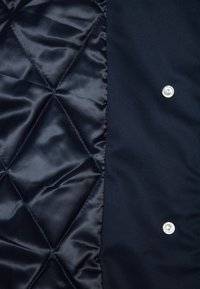 Mörkblå vadderad jackas insida med en slät satinfinish och diamantmönster. Har silverfärgade tryckknappar på vänster sida.