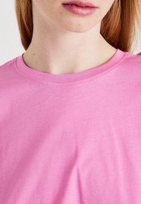 Růžové bavlněné tričko s kulatým výstřihem, které se vyznačuje hladkou texturou a minimem švů. Důraz na rovnoměrnou barvu látky a její splývání.