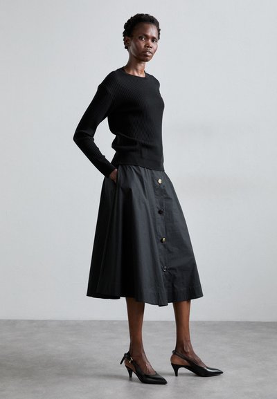 Musträbal riideülikond mustast ribadest, koos musta A-line seelikuga, millel on kuldsed nupud, minimalistlikul hallil taustal; mustad slingback-tossud.