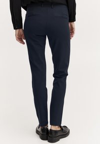 Námornícky modré slim-fit nohavice s hladkým materiálom, dvoma zadnými vreckami a opaskom. Topánky sú lesklé čierne s hrubou podrážkou.