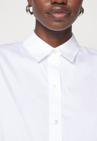 Camicia bianca con colletto, realizzata in tessuto morbido con chiusura a bottoni e bottoni simili a perle; presenta un colletto classico e un design pulito e strutturato.