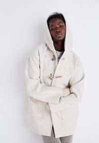 Manteau à capuche en laine beige avec des fermetures à bascule et des poches avant, offrant une texture douce et une coupe décontractée. Porté par-dessus un haut gris.