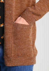 Brązowy sweter na drutach z fakturowaną powierzchnią, ściągaczami na mankietach i przednią kieszenią. Materiał wydaje się miękki i ciepły.