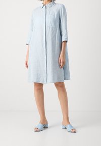 Lichtblauwe gestreepte jurk met lange mouwen en knopenrij aan de voorkant, voorzien van een borstzak. Gepaard met bijpassende blauwe open-toe muiltjes.