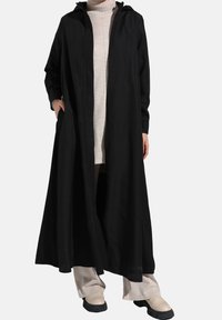 Manteau long noir avec capuche, tissu léger, ouverture à l'avant, et poches. Porté par-dessus un col roulé beige clair et un pantalon crème.