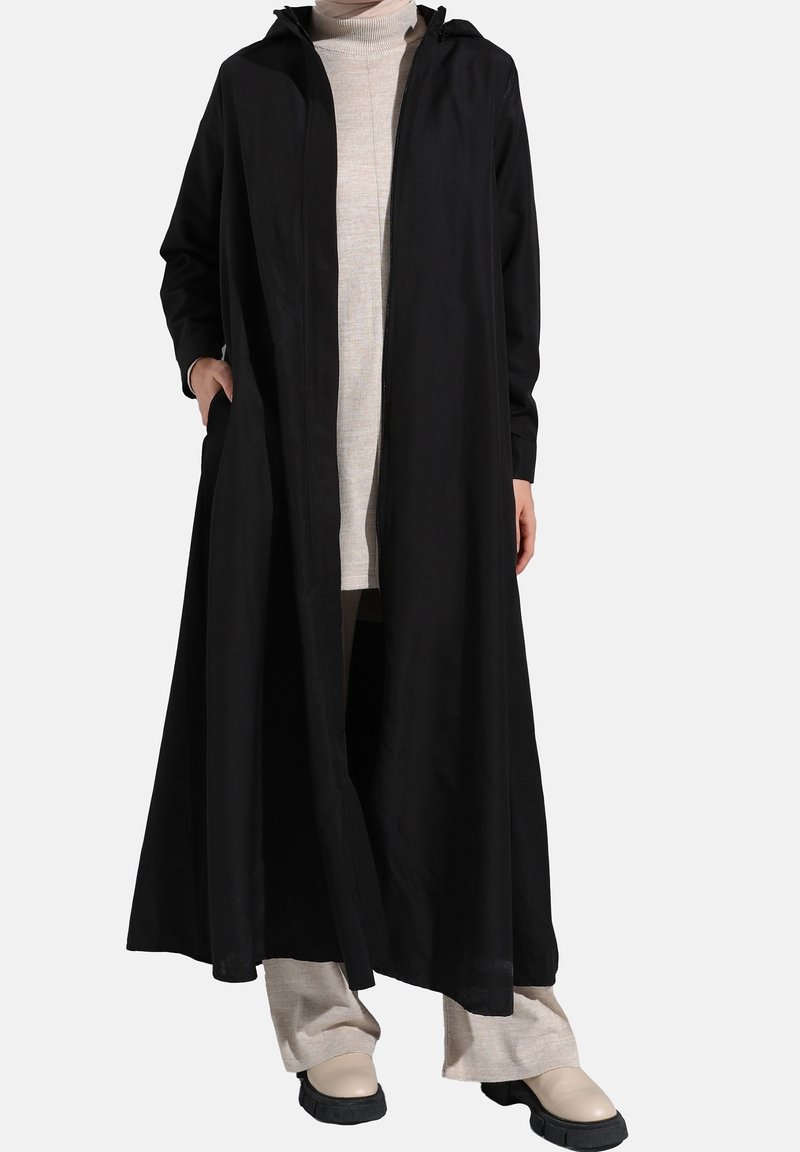 Manteau long noir avec capuche, tissu léger, ouverture à l'avant, et poches. Porté par-dessus un col roulé beige clair et un pantalon crème.